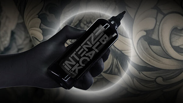 Hand holding a tattoo ink bottle with 'Intenze Black' text against a tattooed background